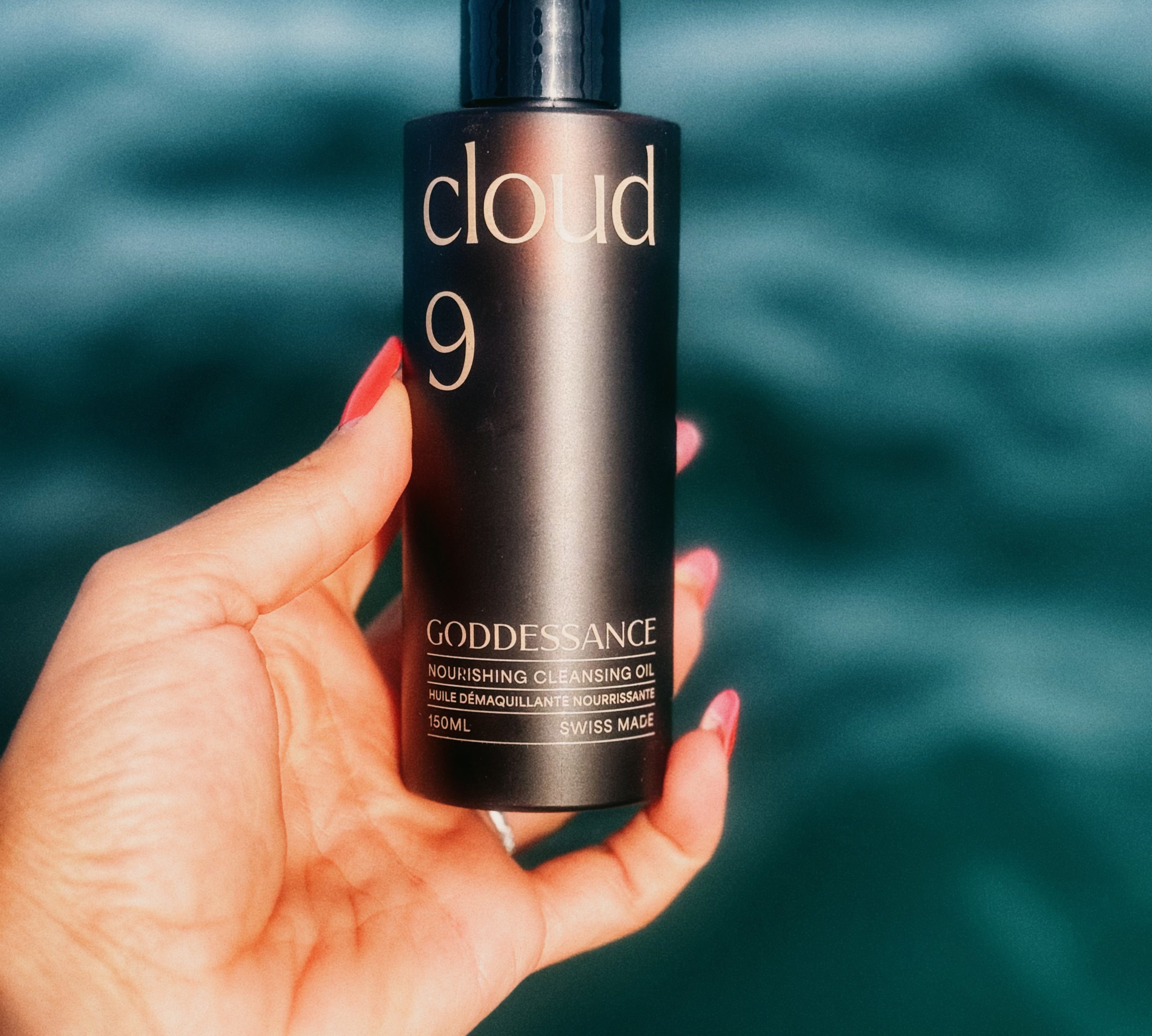 Hand holding a Cloud 9 cleansing oil bottle against a blue sea backdrop