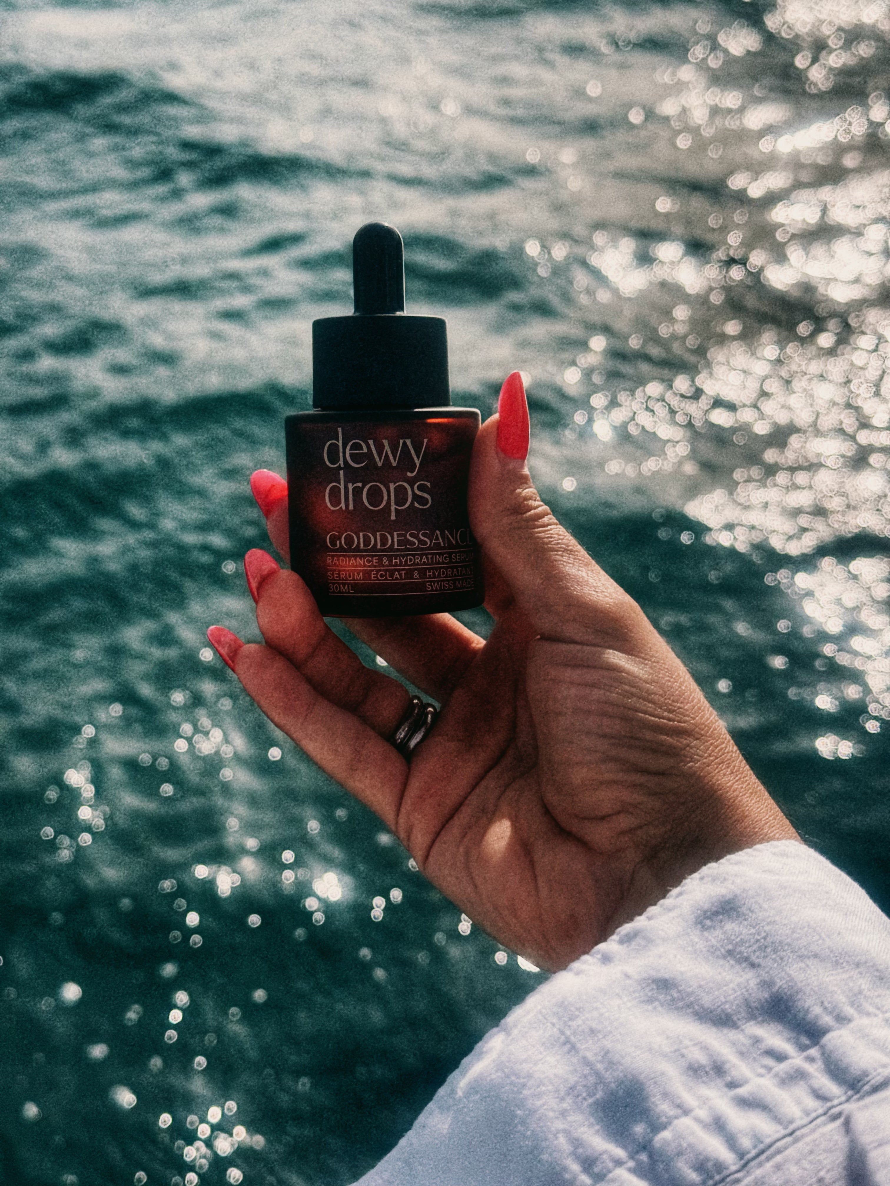 Hand holding a Dewy Drops serum bottle against a sparkling sea